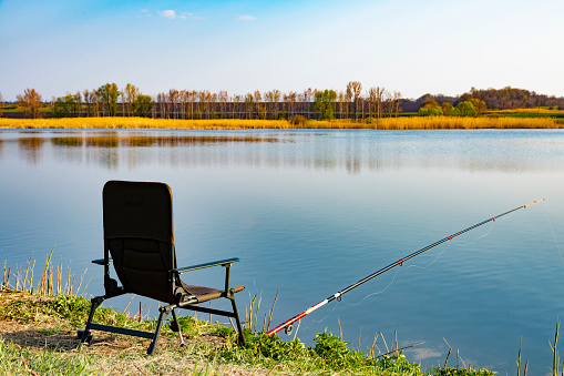 An empty chair and fishing rod on the lake coast in the morning. Green grass near large lake, blue sky, early hours.