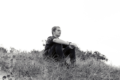 Teenage boy sitting on the hill, black and white, sky background with copy space, horizontal composition