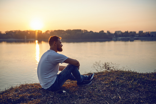 Young handsome bearded caucasian blond man sitting on cliff and looking at sunset. In background is river.