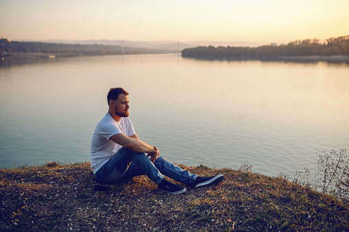 Young handsome bearded caucasian blond man sitting on cliff and looking at sunset. In background is river.