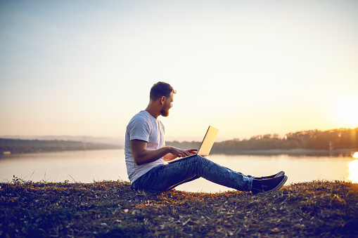 Side view of handsome bearded caucasian blond man sitting on cliff, looking at sunset and typing on laptop. In background is river or lake.