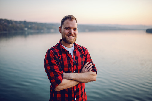 Handsome bearded blond caucasian man standing on cliff with arms crossed and looking at camera. In background is river.