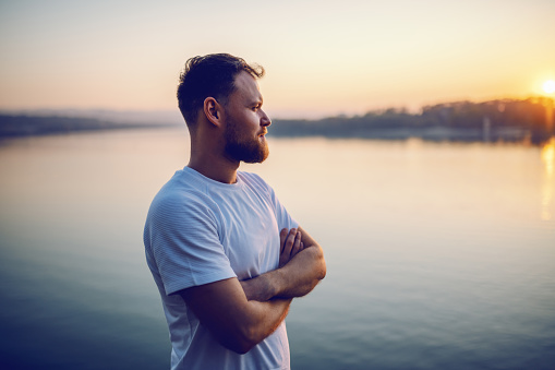 Handsome bearded blond caucasian man standing on cliff with arms crossed and looking at beautiful sunset. In background is river.