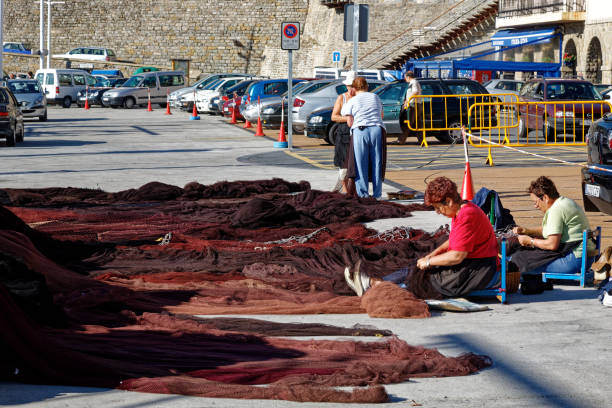 Ladies on the quayside sitting down repairing A large pile of fishing nets with white floats in the sun.