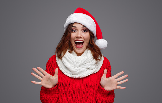 Excited eccentric female in red Santa hat and colorful knitwear screaming with open palms looking at camera