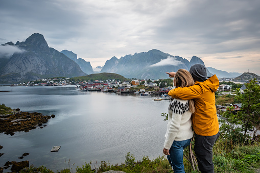 Couple visiting the fishing village of Reine in the Lofoten Islands with mountain range and cloudy sky in background during summer.