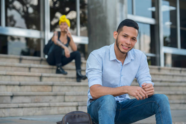 Millennial man in the street while a woman is with his cell phone behind Latin American millennial man between the ages of 20-30 who has tattoos on the street while a woman is with his cell phone behind latin script stock pictures, royalty-free photos & images