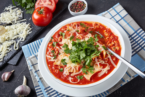 close-up of chicken lasagna soup in a white bowl on a grey concrete table, american italian cuisine. ingredients at the background, view from above
