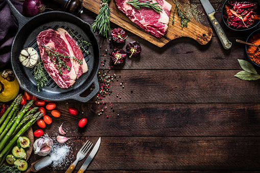 Top view of an iron grill with a beef steak, aromatic herbs and garlic surrounded by vegetables like tomatoes, asparagus and roasted Brussel sprouts, salt and pepper, aromatic herbs like thyme and rosemary, a kitchen knife and a wooden cutting board with a seasoned beef steak on top on a dark brown rustic wooden table. Objects are at the top and at the left of the image leaving a useful copy space for a text or a logo at the lower right corner on the backdrop. Low key DSLR photo taken with Canon EOS 6D Mark II and Canon EF 24-105 mm f/4L