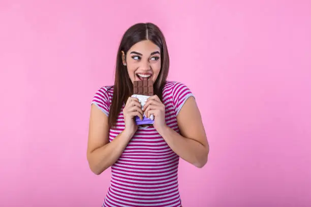 Photo of Portrait of a pretty young girl eating chocolate bar isolated over pink background. Satisfied pretty girl biting chocolate bar