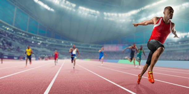 Champion Athlete Wins Sprint Race Competition In Indoor Track Event Wide angle, shallow depth of field image of a professional male track athlete sprinting over the finish line of a race with his arms outstretched in victory with composed expression. The athlete is competing in an indoor track sprint event in a generic arena, and finishes ahead of his competitors. sprint stock pictures, royalty-free photos & images