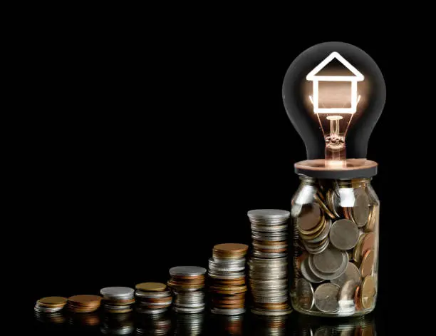 Group of coin rows, glass jar and light bulb with shining fiber in a shape of house icon isolated on black background. Concept of property and house investment, mortgage and money save.