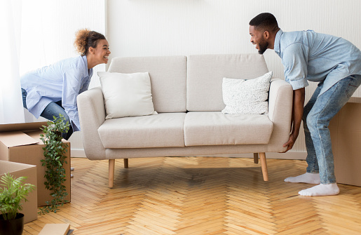Moving Concept. Cheerful Afro Spouses Placing Couch Furnishing Empty Room In New House After Relocation