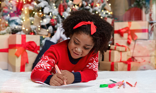Letter to Santa. Cute little black girl lying on floor and writing wish list of presents for Christmas in decorated room