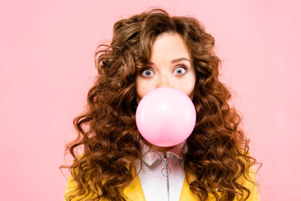 ピンクに隔離されたバブルチューインガムを持つ魅力的な驚いたカーリーガール - chewing gum ストックフォトと画像