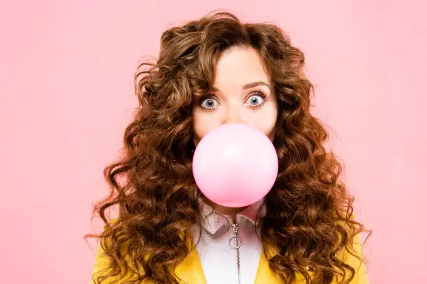 Photo of attractive surprised curly girl with bubble chewing gum, isolated on pink