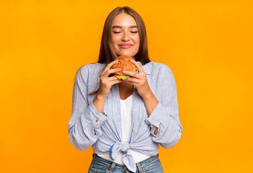 Junk Food. Cheerful Woman Smelling Burger Standing Over Yellow Background. Studio Shot