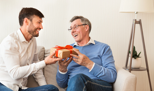 Mature Son Congratulating Elderly Father Giving Him Birthday Gift Sitting On Sofa At Home. Selective Focus