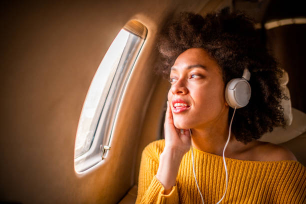 jeune femme à la mode s'asseyant sur un avion privé et regardant par une fenêtre tout en écoutant la musique par des écouteurs - status symbol audio photos et images de collection