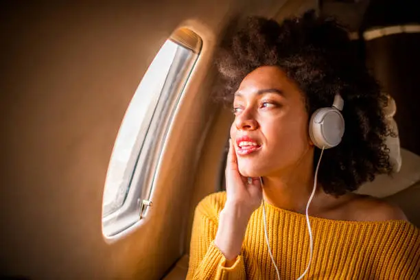 Young fashionable woman sitting in a private jet and listening to music through the headphones. She is looking through the window.