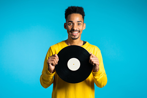 Young handsome african american man in yellow wear holding vinyl records at blue background. Retro style music concept