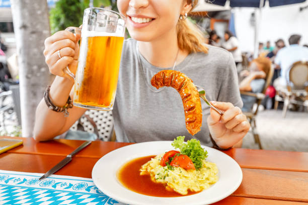 uma menina asiática feliz bebe uma caneca da cerveja do lager em um biergarten bávaro tradicional e petiscos em uma salsicha suculenta e em uma salada de batata. conceito alemão da culinária da guloseima - german culture salad prepared potato bratwurst - fotografias e filmes do acervo