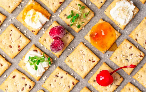 Homemade freshly baked crackers with flax and sesame seeds with various serving options are laid out on baking paper, top view. Crackers with seeds, ricotta, jam, berries and microgreens, flat lay.
