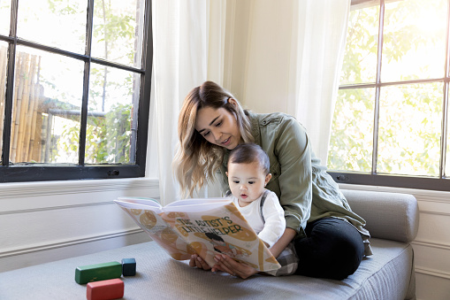 Adorable baby girl looks attentively at a book as her mid adult mom reads to her.