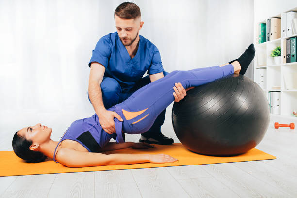 istock Physical therapist helping his patient doing exercise. Woman laying on mat and doing exercise with pilates ball 1187859579