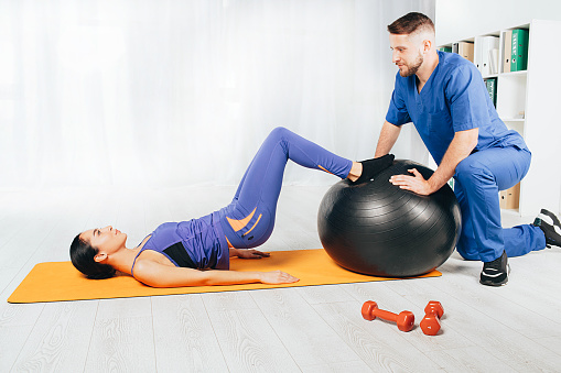 Physical therapist working with a young woman. She lying on exercise mat and doing exercise with pilates ball