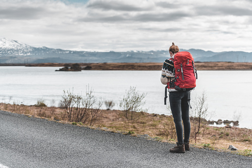 The woman with a red rucksack stands on a lake shore, Þjórsárdalsvegu, Southern Region of Iceland.