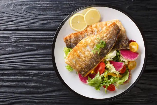 Photo of Golden grilled sea bass fillet with lemon and healthy vegetable salad close-up on a plate. Horizontal top view