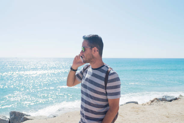 istock Young man in t-shirt standing on seashore while using a mobile phone 1187876256