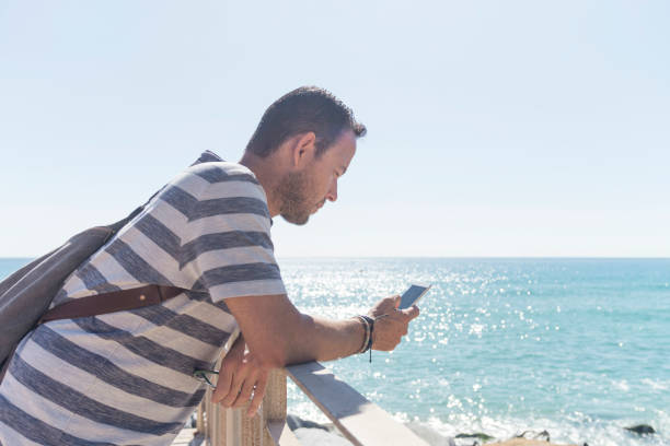 istock Side view of a young man in t-shirt while using mobile phone on seashore in a sunny day 1187876269