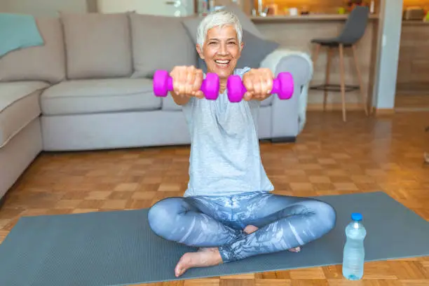 Portrait of mature fitness woman lifting dumbbells. Portrait of happy senior woman holding dumbbell at home. Active senior woman exercising with dumbbell at home