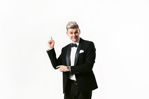 Portrait of young smiling handsome man in tuxedo stylish black suit, studio shot isolated on white background. Showman or toastmaster in jacket with bowtie. He points at copy space by hand