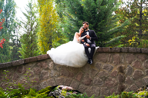 Bride and groom romantic bridge over river. Beautiful young bride with elegant hairdo in white wedding dress with shoes sitting on stone bridge parapet