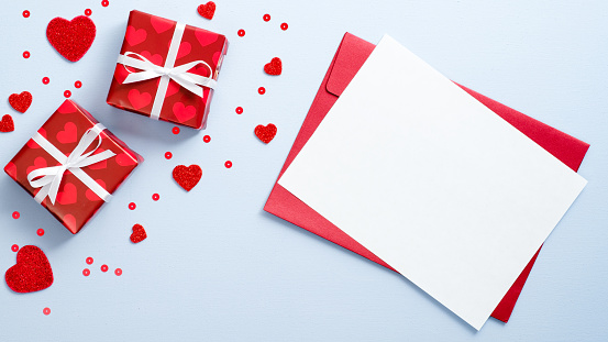 Red envelope with blank paper card and gift boxes decorated hearts on blue background. Romantic letter concept. Happy Valentine Day flat lay composition.