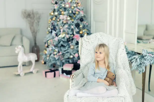 cute adorable little girlsitting in chair near christmas tree in cozy living room looking , dreaming and waiting xmas wonder. Portrait of blond kids.