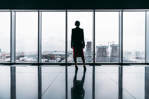 Rearview of a female entrepreneur on the top of a modern business office skyscraper standing near big panoramic windows and pensively looking at cityscape and construction site; floor with reflections