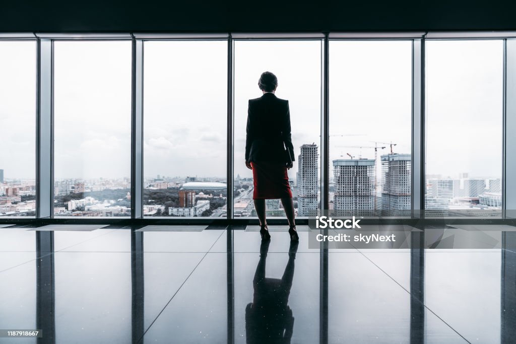 A silhouette of a businesswoman Rearview of a female entrepreneur on the top of a modern business office skyscraper standing near big panoramic windows and pensively looking at cityscape and construction site; floor with reflections Women Stock Photo