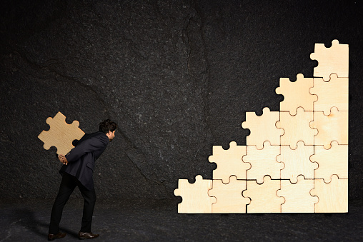 A businessman is carrying a piece of puzzle to an unfinished puzzle on a black textured wall
