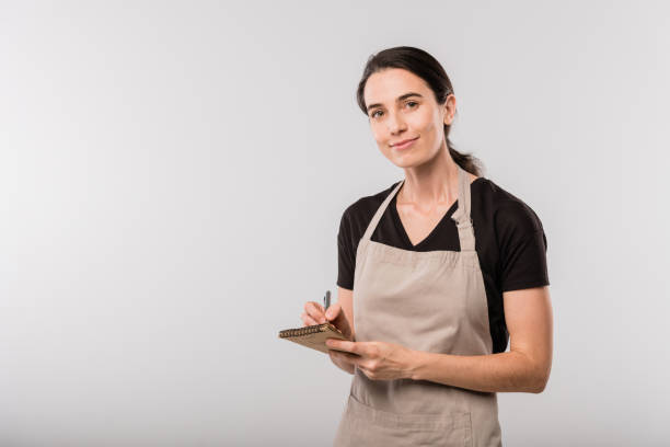 istock Pretty young brunette waitress in apron making notes about order in notepad 1187980439