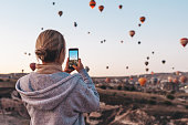 Woman taking photo on beautiful landscape and balloons in Cappadocia with mobile camera, sunrise time.