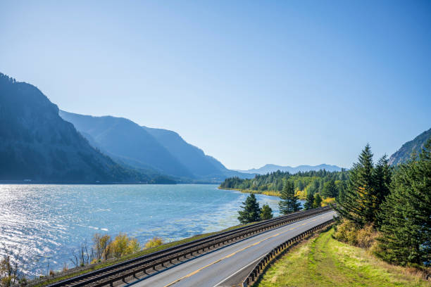 Railway track and vehicles road along the Columbia River in Columbia Gorge A railroad track next to a highway road with lane dividing opposite traffic runs along the scenic Columbia River bank with green trees and rocky mountains in Columbia Gorge area northwest stock pictures, royalty-free photos & images