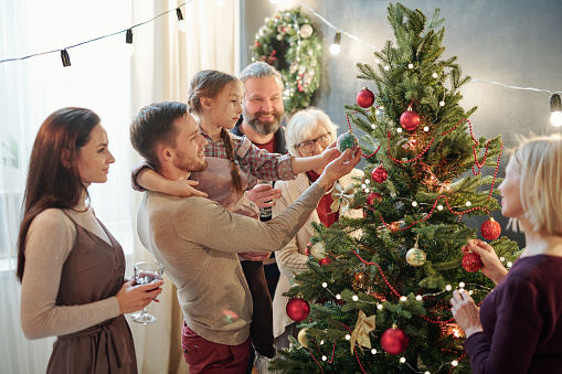 Young man with his little daughter and other family members standing by Christmas tree at home while preparing for holiday