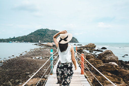 Summer travel vacation concept, Happy traveler asian woman with white dress and hat relax and walk on wooden bridge in tropical beach, Thailand