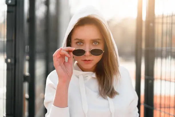 Outdoor close up portrait of young beautiful woman with long hair in sunglasses, dressed in a white hoodie sweater, near the sportsground. youth culture summer pastime
