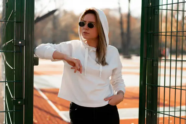 Outdoor close up portrait of young beautiful woman with long hair in sunglasses, dressed in a white hoodie sweater, near the sportsground. youth culture summer pastime