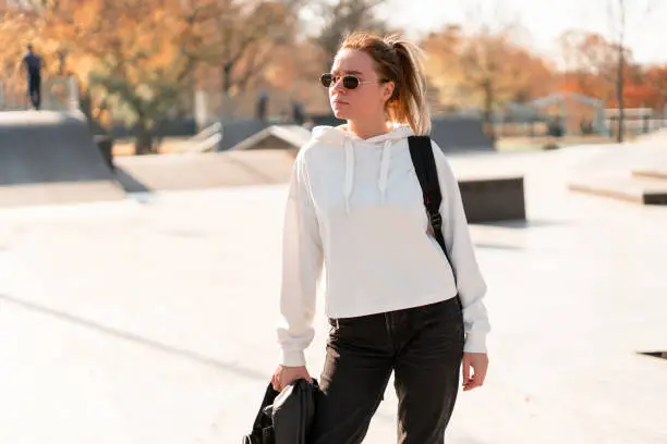 Outdoor portrait of young beautiful woman with a ponytail and sunglasses, with a backpack on his shoulders, dressed in a white sweater, near the sportsground. White hoodie